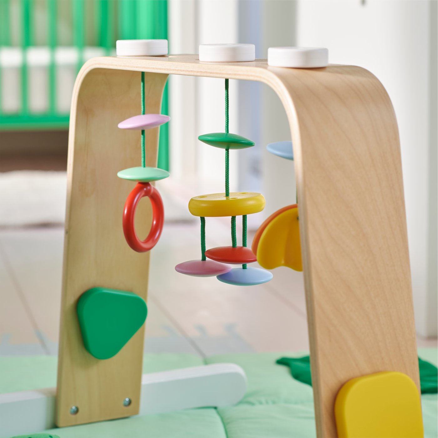 A multicolour LEKA baby gym with bright coloured hanging rings and shapes on top of a green playmat in a baby’s bedroom.
