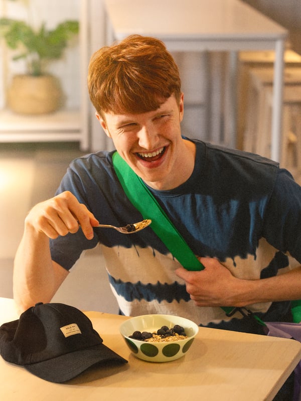 Eine Person sitzt an einem Tisch und hält einen Löffel Joghurt mit Blaubeeren in der Hand, neben einer Schüssel Joghurt und einer schwarzen Kappe.