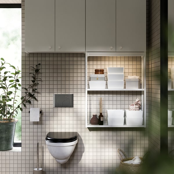 A white ENHET open storage with two shelves hangs in a softly lit bathroom. It’s filled with white boxes and dark bottles.