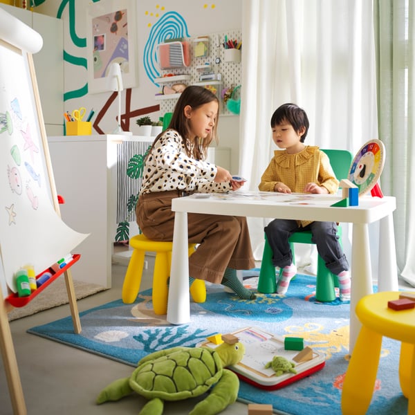 A child sits on a green MAMMUT children’s chair at a table with an adult who is sitting on a yellow MAMMUT children’s stool.
