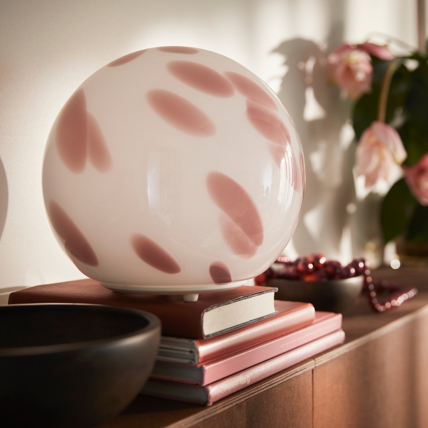 A close-up of a pink FADO table lamp made of glass that sits on stacked books beside a brown bowl and a plant.