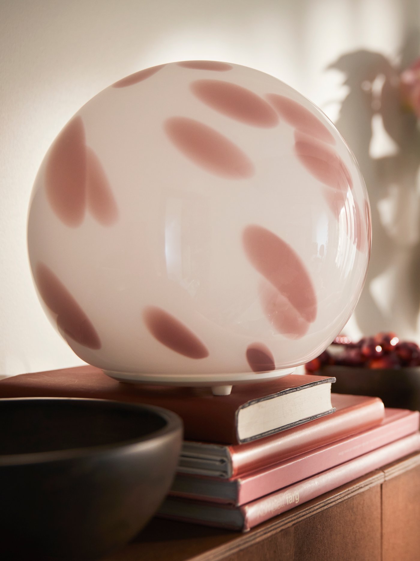 Een close‑up van een roze FADO tafellamp van glas die op een stapel boeken staat, naast een bruine schaal en een plant.