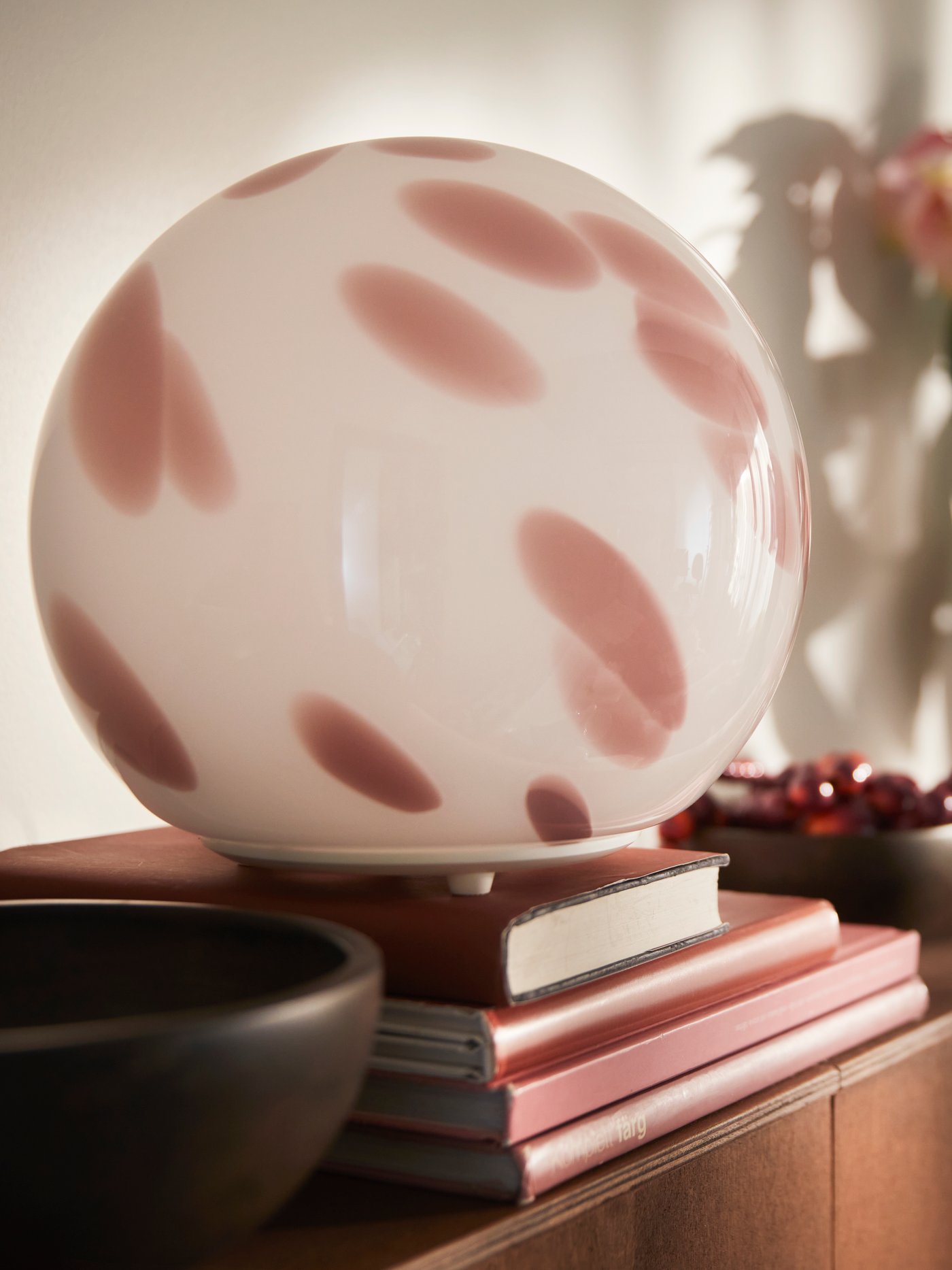 Zbliżenie na różową lampę stołową FADO wykonaną ze szkła, ustawioną na stosie książek obok brązowej miski i rośliny.