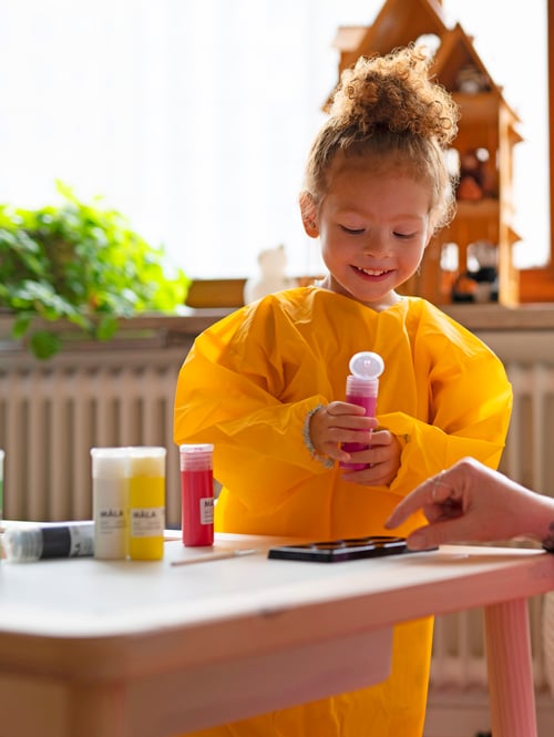 A child wearing a yellow MÅLA apron pouring paint into a plastic container.