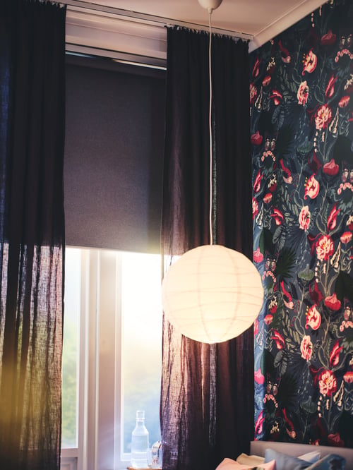 Schlafbereich mit einem halb geschlossenen SKOGSKLÖVER Rollo, einem REGOLIT Hängeleuchtenschirm, DYTÅG Gardinenschals und FILODENDRON Meterware an der Wand hinter dem Bett.