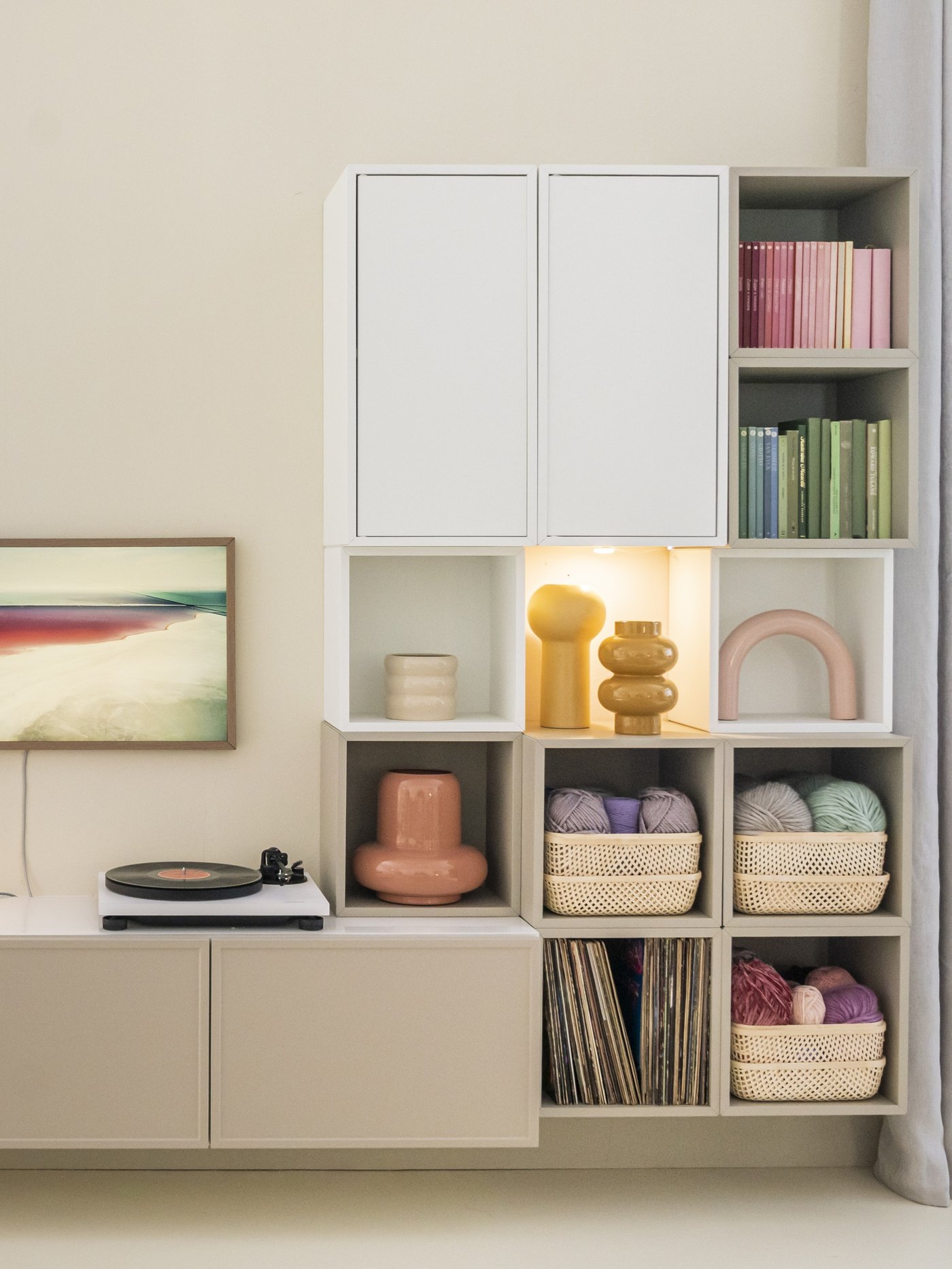 Composizione EKET bianco e beige a parete con vani chiusi e a giorno, usato per libri, cestini e oggetti decorativi in un soggiorno moderno.