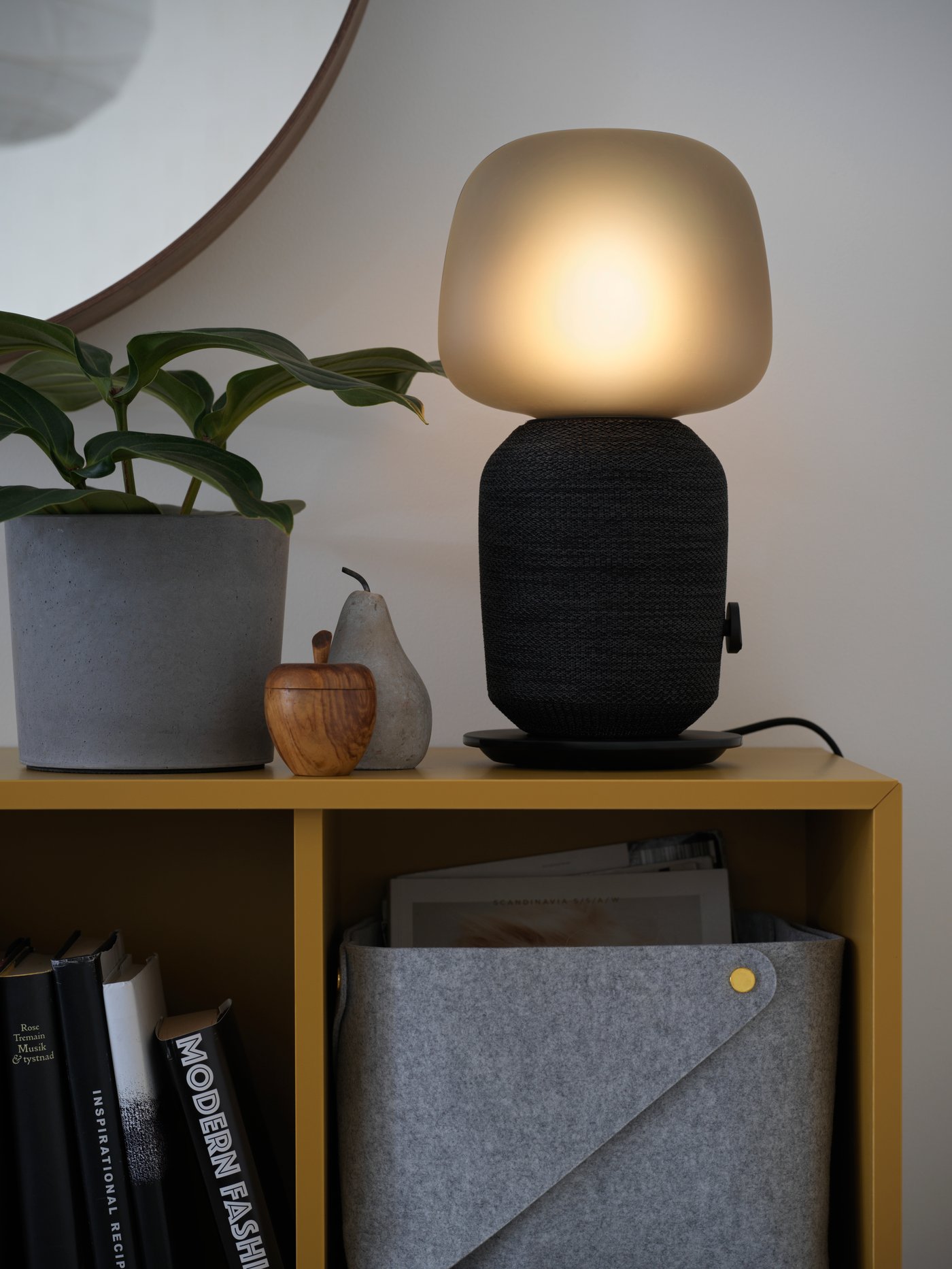 A black SYMFONISK table lamp with WiFi speaker, next to a houseplant and ornaments, on top of EKET storage cabinets.
