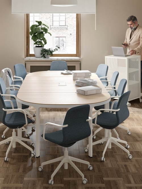Workspace with multiple office chairs with armrests surrounding an oval conference table, and a man sitting at a storage unit.