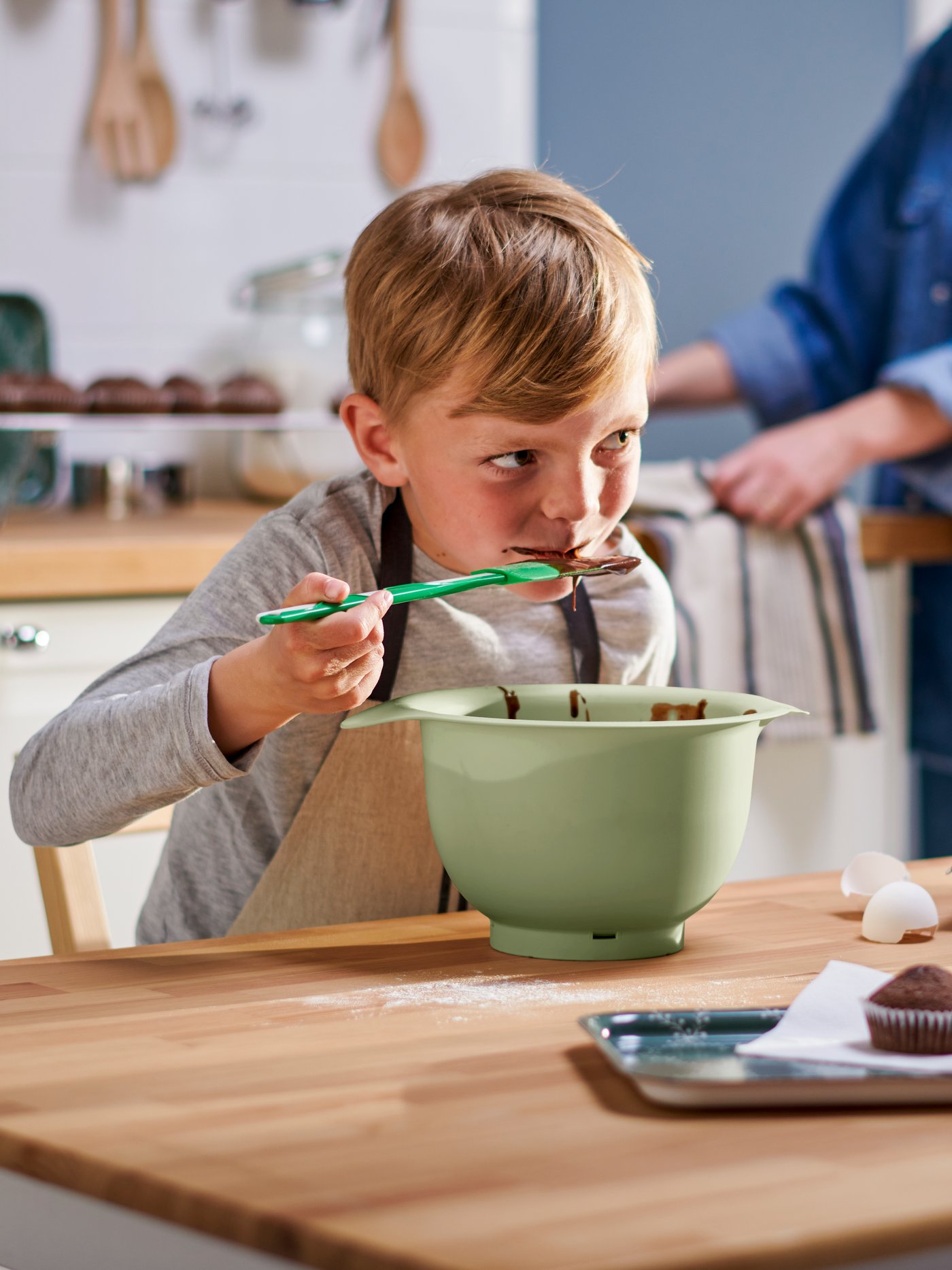 Ein Kind in einer MARIATHERES Kinderschürze probiert den Teig eines Schokoladenkuchens, der sich in einer grünen Rührschüssel befindet, mit einem GUBBRÖRA Teigschaber in Grün/Weiss.