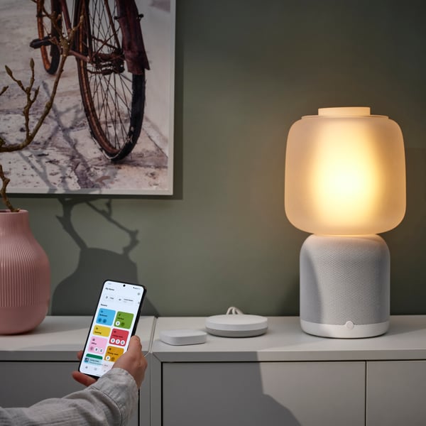 Ein weisser SYMFONISK Leuchtenfuss mit WiFi-Speaker und einem Glasschirm steht auf einem Schrank. Eine Hand zeigt mit einem Handy darauf.