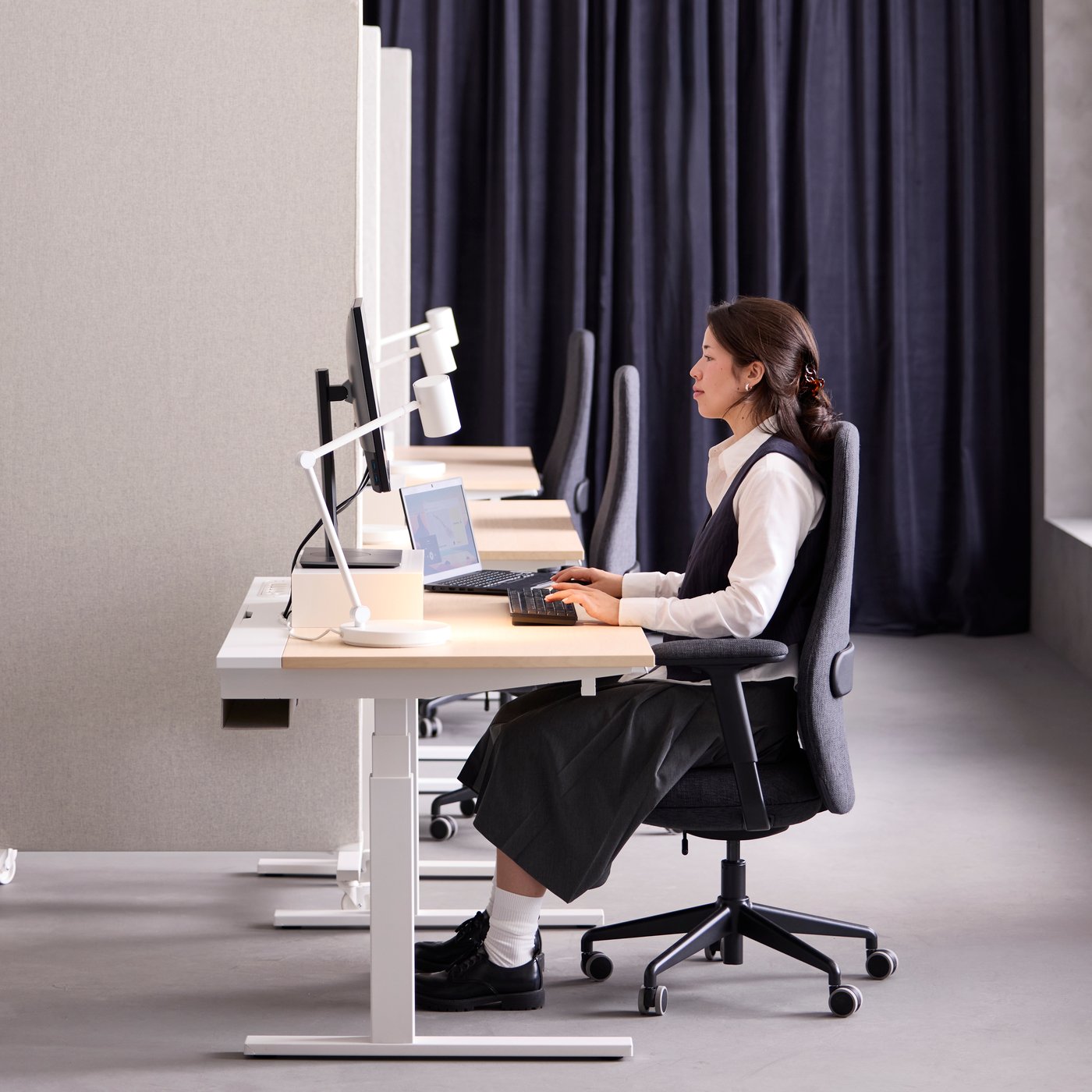 Office scene with a person using a white MITTZON birch veneer sit/stand desk on low mode, sitting by a computer setup.
