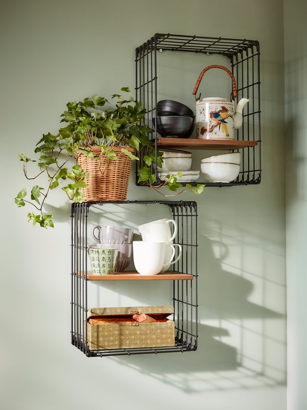 Two black wire wall shelves with teacups, a ceramic tea pot, a box with tea and a plant in a wicker pot.