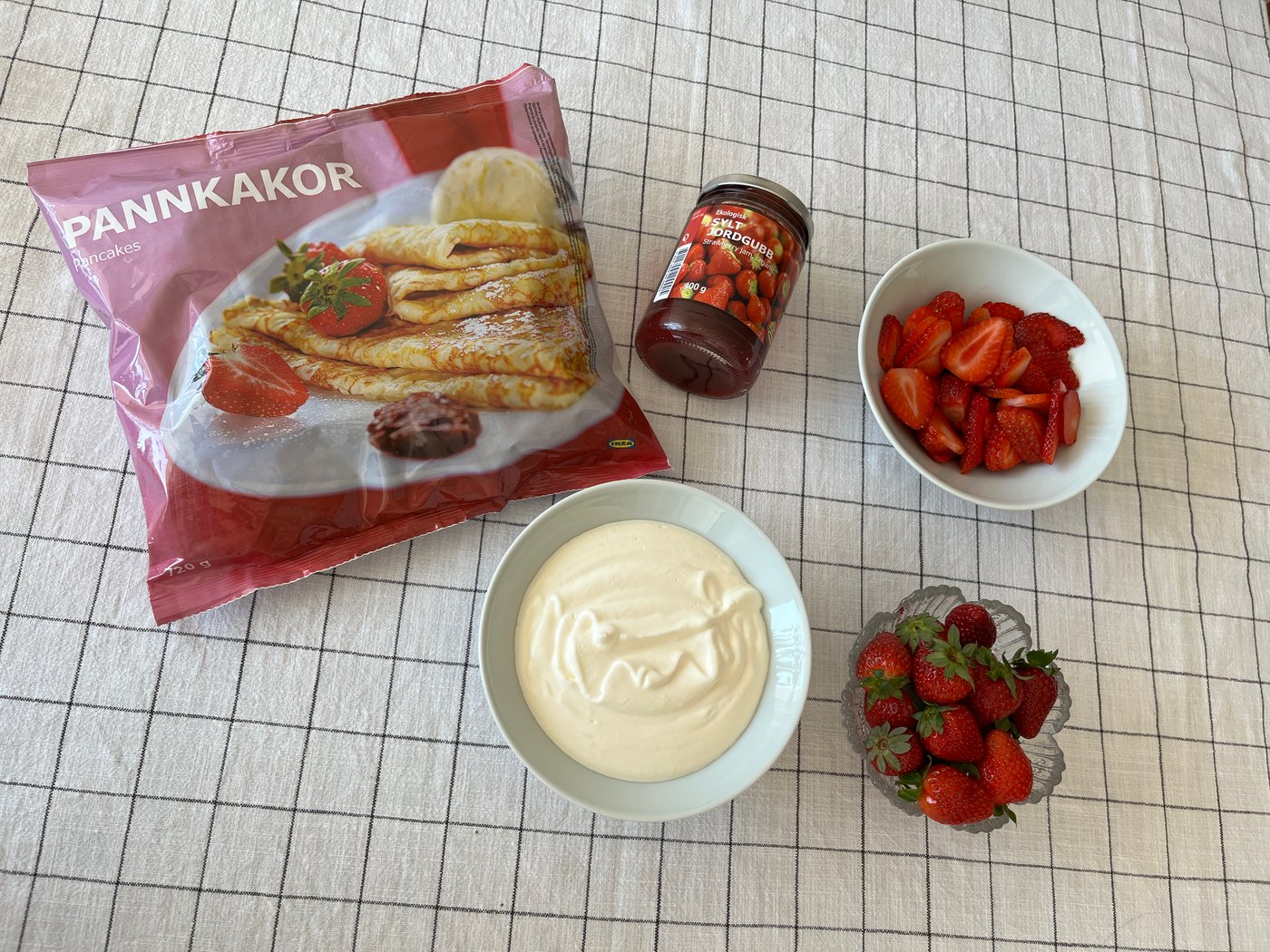 På ett bord med rutig duk syns en tallrik med en pannkakstårta, färgglatt randigt porslin, en skål med jordgubbar och en vas med blommor i.