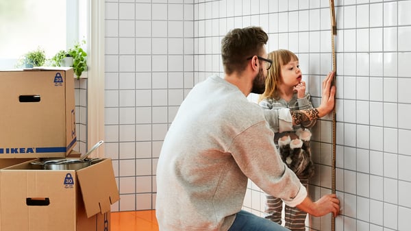 A person measures a tiled kitchen wall, with IKEA flat‑pack boxes stacked nearby.