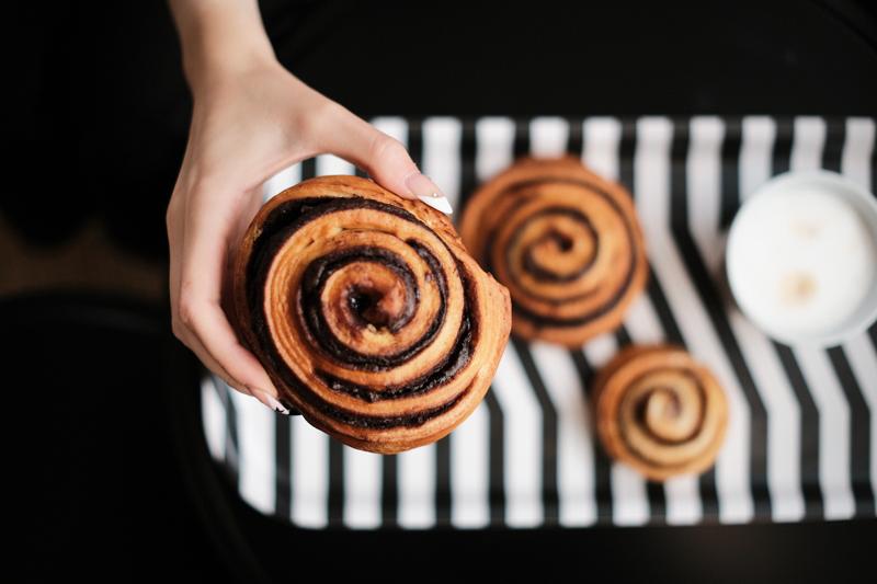 Eine Hand hält eine Schokoladenschnecke. Darunter sind zwei weitere Schnecken auf einem schwarz-weiß gestreiften Tablett zu sehen,