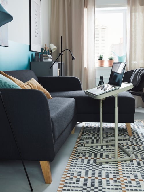 A LININÄS sofa by a sunny window, with beige and turquoise cushions on it and a laptop stand in front of it holding a laptop.