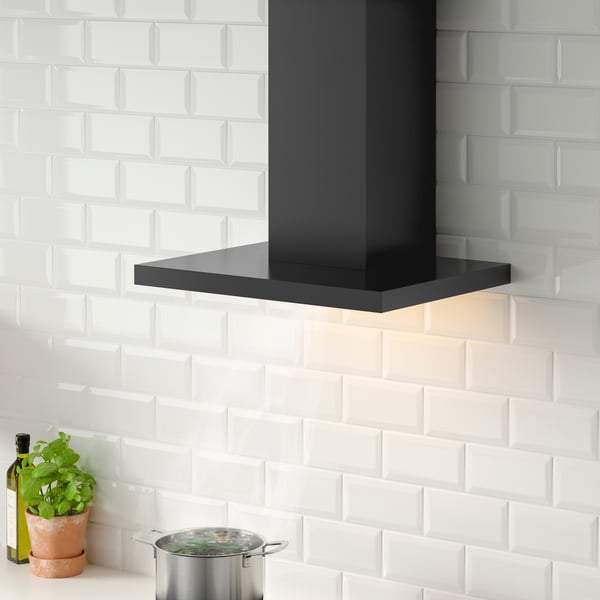 Eine MATÄLSKARE Dunstabzugshaube für die Wandmontage in Schwarz hängt an einer weißen Küchenwand. Darunter befindet sich ein Topf aus Edelstahl.