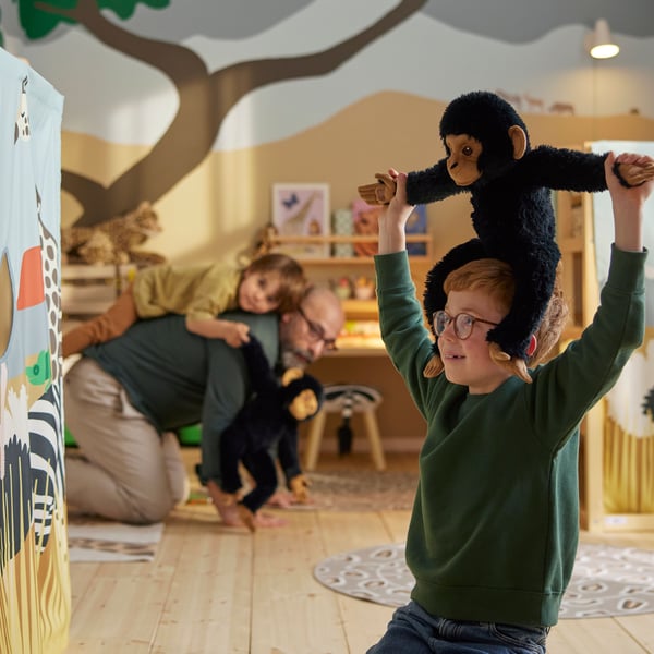 A child sits on their knees in a savannah themed room while carrying a SANDLÖPARE soft toy chimpanzee on their shoulders.
