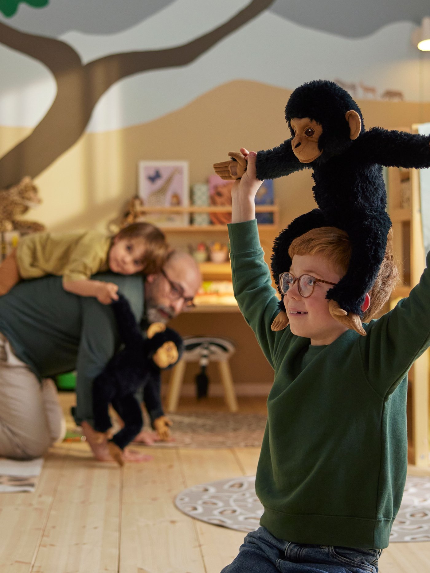 A child sits on their knees in a savannah themed room while carrying a SANDLÖPARE soft toy chimpanzee on their shoulders.