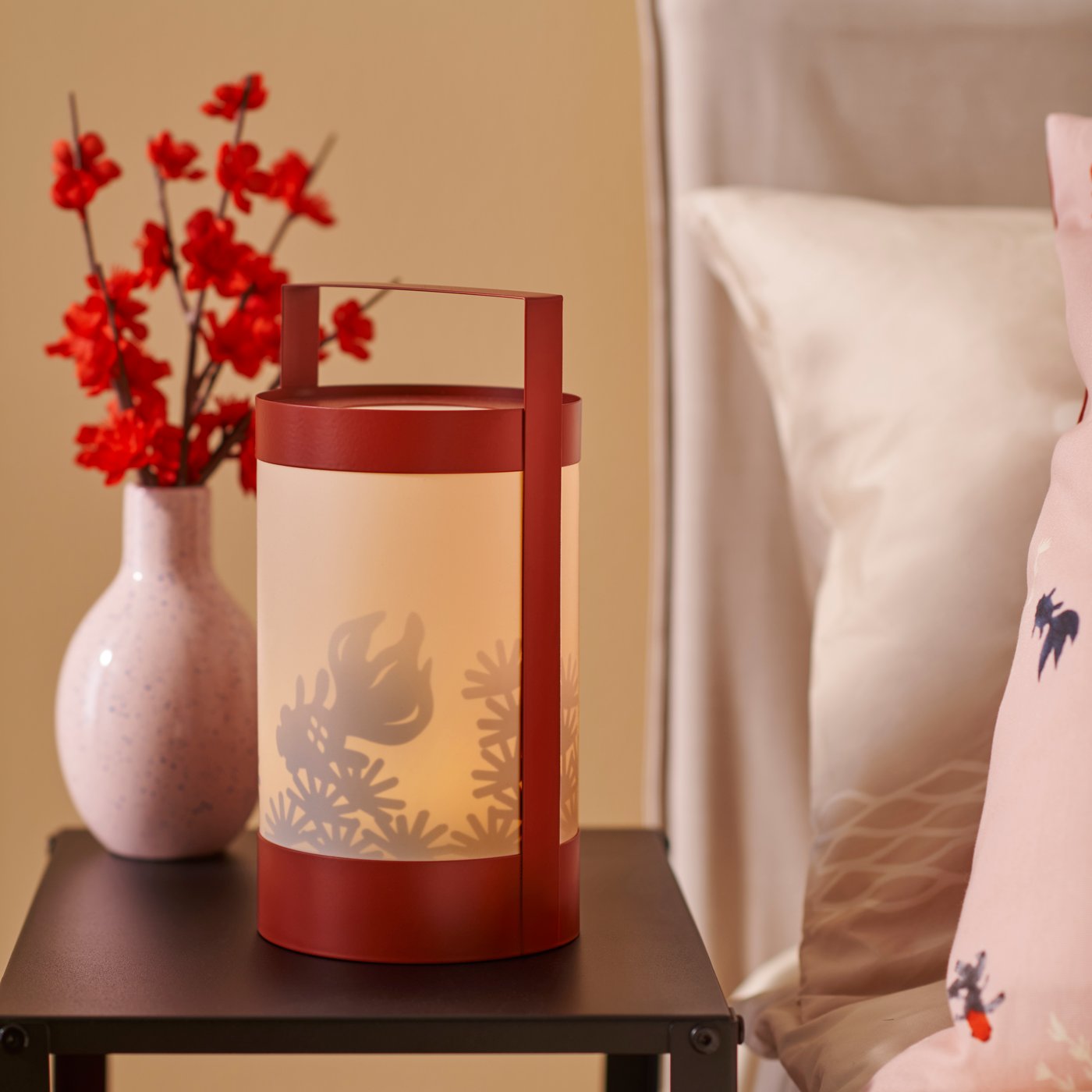 A FÖSSTA LED table decoration is placed on a dark table between a bed and a vase containing red flowers.