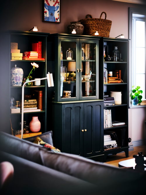 Un salon avec une armoire bleue à portes vitrées, une bibliothèque, un vase rose, des éclairages à DEL et une lampe de lecture sur pied.