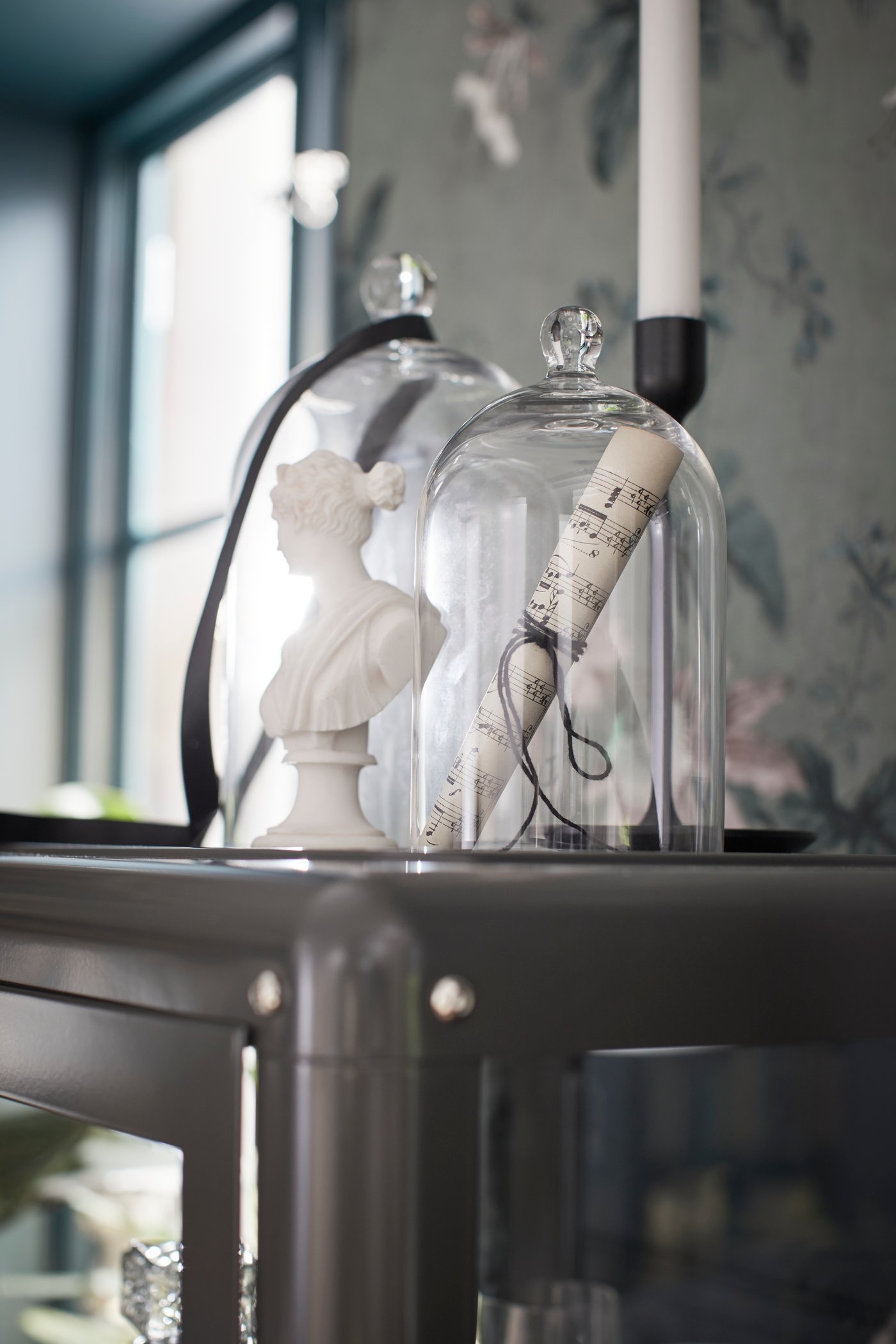 Ein Kerzenhalter und zwei MORGONTIDIG Glasglocken auf einem Schrank. Die Dekorationen unter den Glasglocken teilen ein musisches Thema.