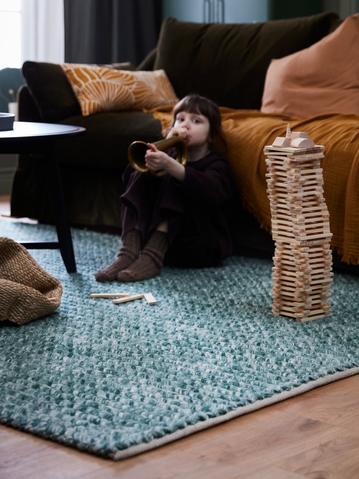 Ein Kind spielt mit einem Holzstapelspiel auf einem dunkelgrünen AVSKILDRA Wollteppich. Dabei lehnt es sich gegen ein FÄRLÖV Sofa.