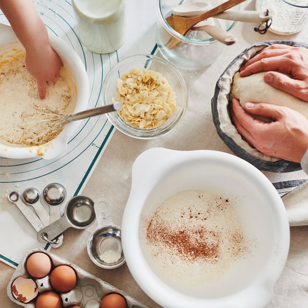 Vista dall'alto di impasti che vengono preparati in diverse ciotole VISPAD. Ci sono 2 persone che stanno cucinando e attorno accessori da cucina in vasi CYLINDER e alimenti. come farina e uova.