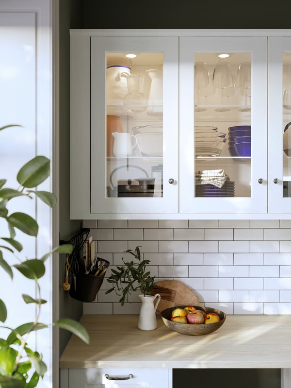 Glass cabinets with white ENKÖPING frames have kitchenware stacked on shelves, illuminated by MITTLED spots on the inside.