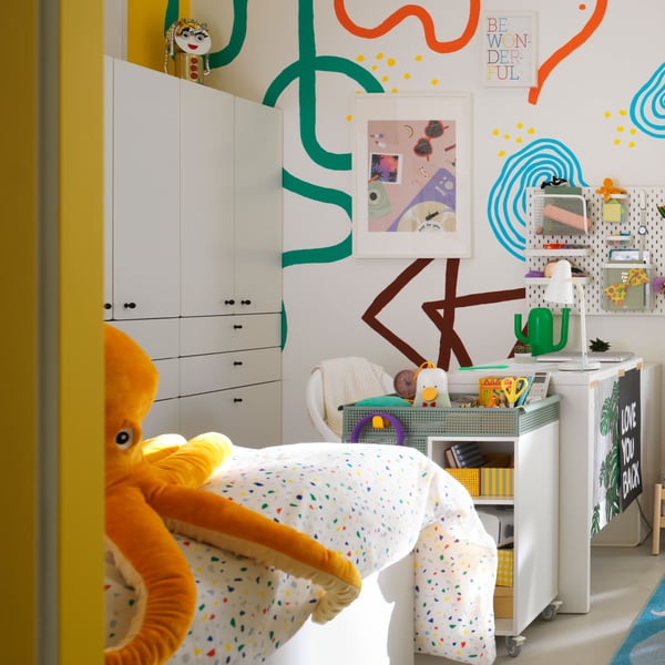 Une chambre d’enfants colorée et dynamique à l’atmosphère ludique. Sur le côté gauche, une grande pieuvre en peluche orange est posée sur une surface à pois. Au centre, unité de rangement blanche renfermant divers jouets et livres et sur laquelle se trouvent des objets décoratifs ; à côté, image murale abstraite en orange, bleu, vert et brun. Sur la droite, table enfant entourée de jouets, dont des blocs de construction et un petit chevalet. Le mur du fond présente d’autres œuvres abstraites près d’une fenêtre habillée de voilages laissant entrer la lumière naturelle.