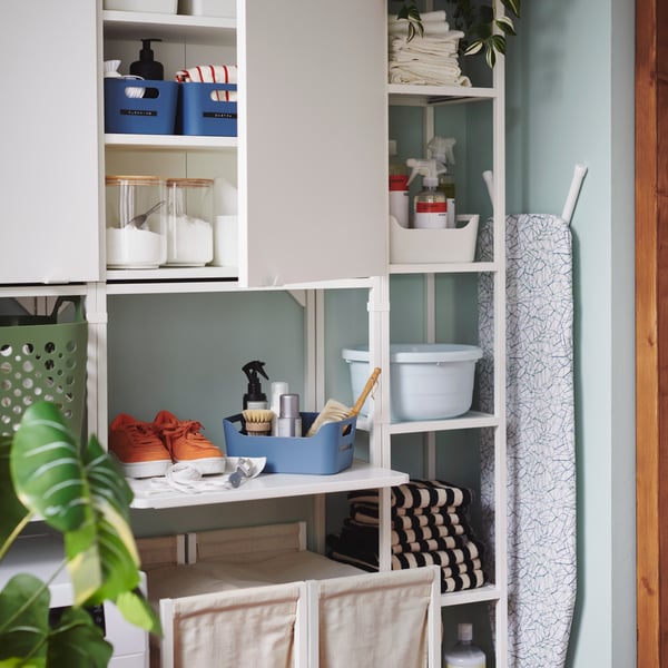 A grey-blue UPPDATERA box holds cleaning tools by shoes on a deep wall shelf above a washing machine in an enclosed balcony.