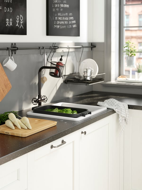 A black colander holding vegetables resting over a white HAVSEN sink. A chopping board is on the countertop beside it.