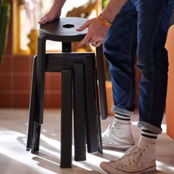 A person is standing by three dark grey IBBARBO stools stacked on each other, holding a fourth one slightly above the rest.