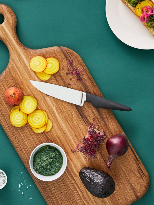 A VÖRDA utility knife is placed on an ARTISTISK chopping board in oak with a variety of vegetables, some of them sliced.