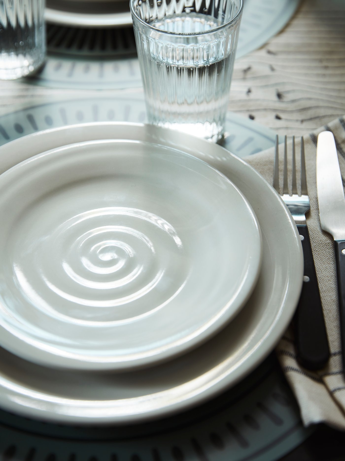 A SANDSKÄDDA side plate sits on top of a SANDSKÄDDA plate next to a VARDAGEN glass and a knife and fork on a table.