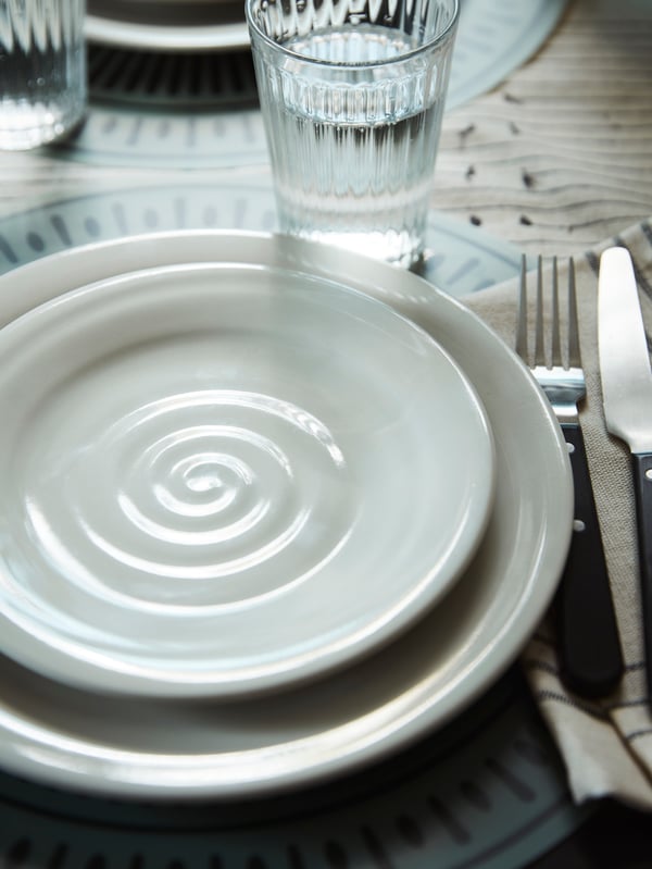 A SANDSKÄDDA side plate sits on top of a SANDSKÄDDA plate next to a VARDAGEN glass and a knife and fork on a table.