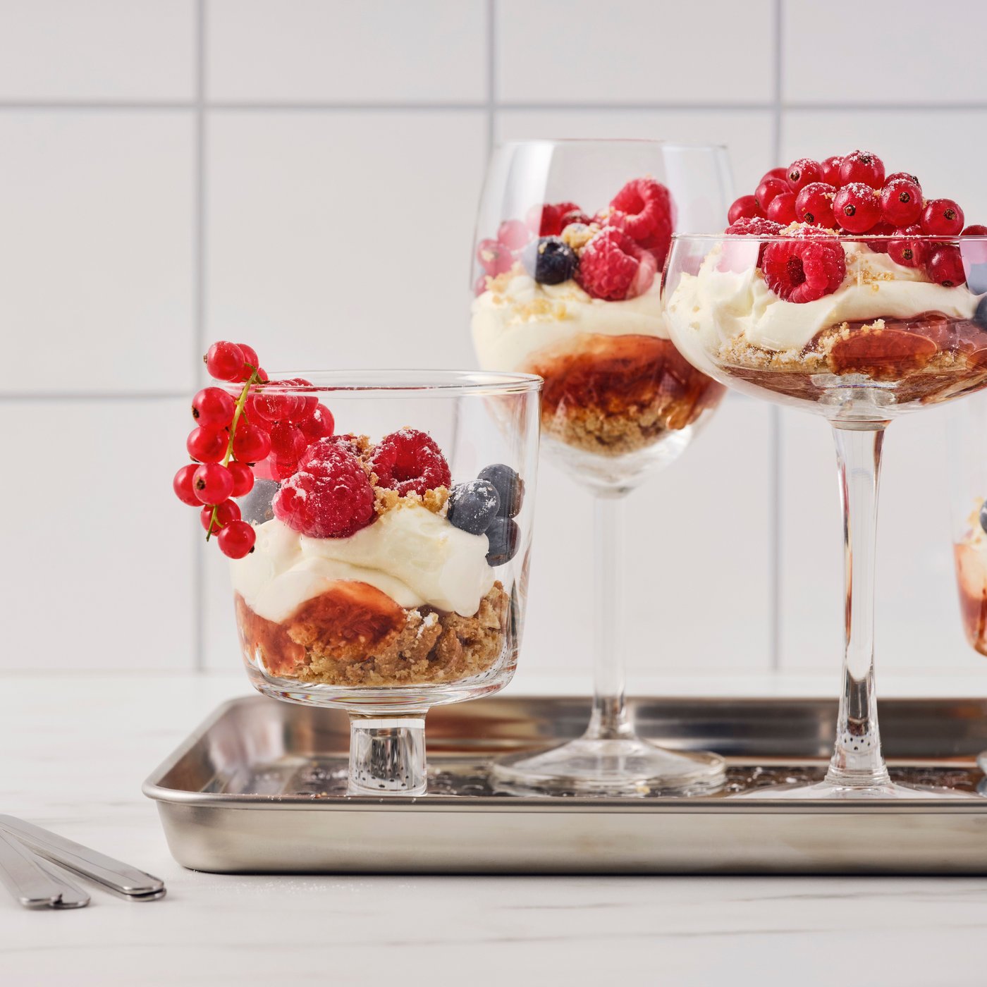Several glasses of different shapes, with oat biscuit and raspberry jam cheesecake inside, are placed on a metal tray.