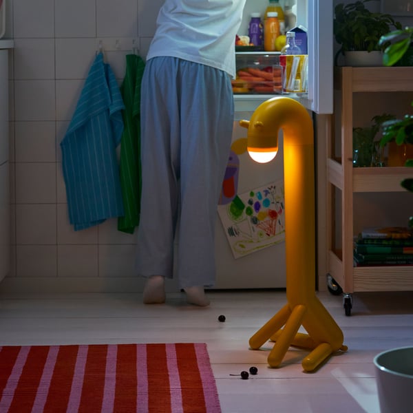 In a dark kitchen, a GREJSIMOJS giraffe LED floor lamp provides light while a person in pyjamas searches the refrigerator.