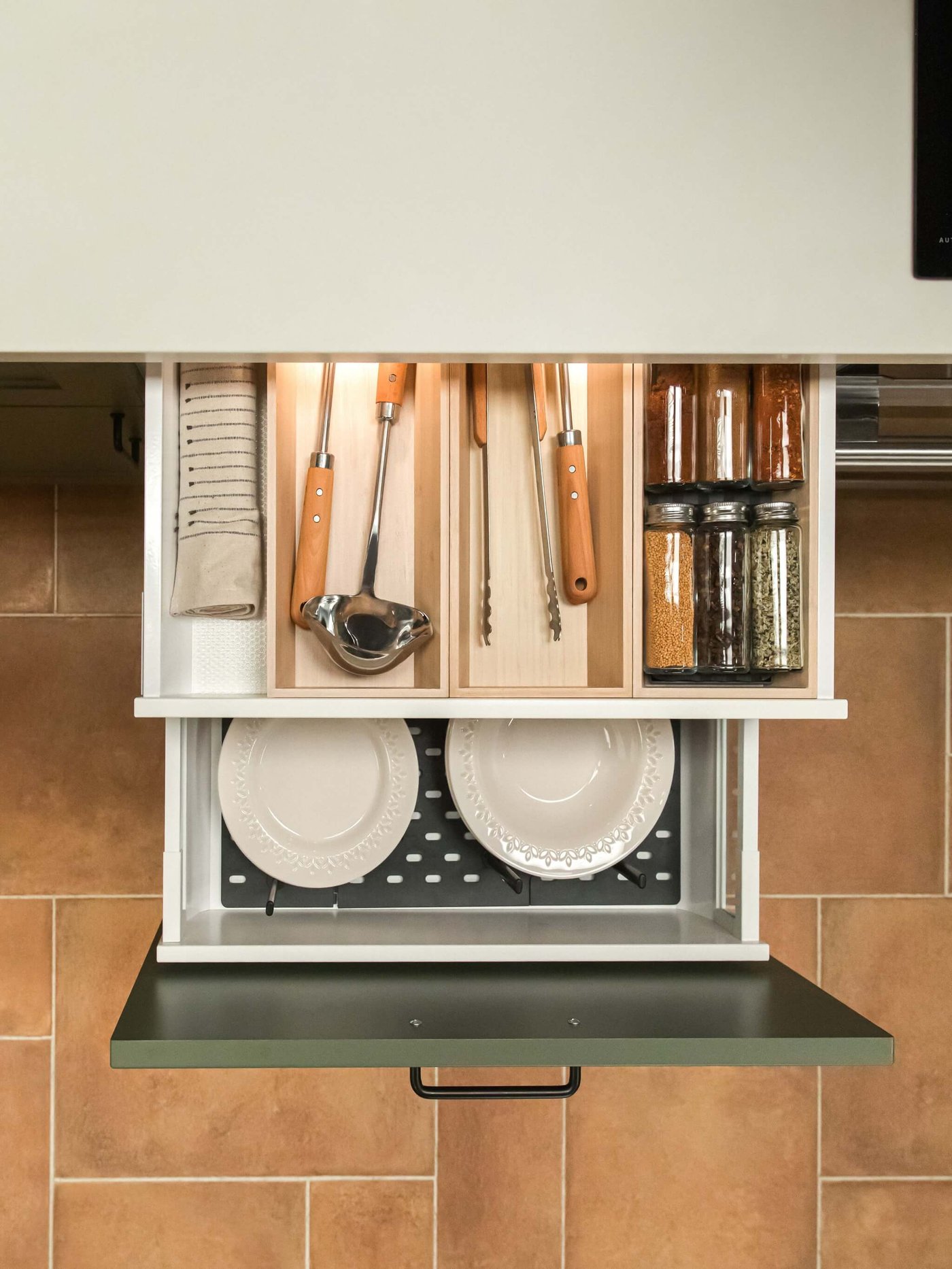 Organized kitchen cabinet with wooden compartments holding utensils, spice jars, and towels; plates stored neatly below.