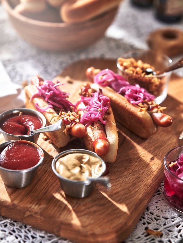 Hot Dogs mit roten Zwiebeln, Röstzwiebeln, Ketchup, Senf auf Holzbrett.