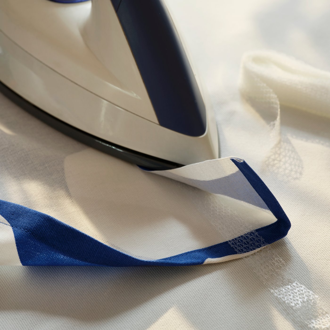 An iron pressing on a SY iron-on hemming strip placed in the folded hem of blue-and-white RAKLOSTA pre-cut fabric.