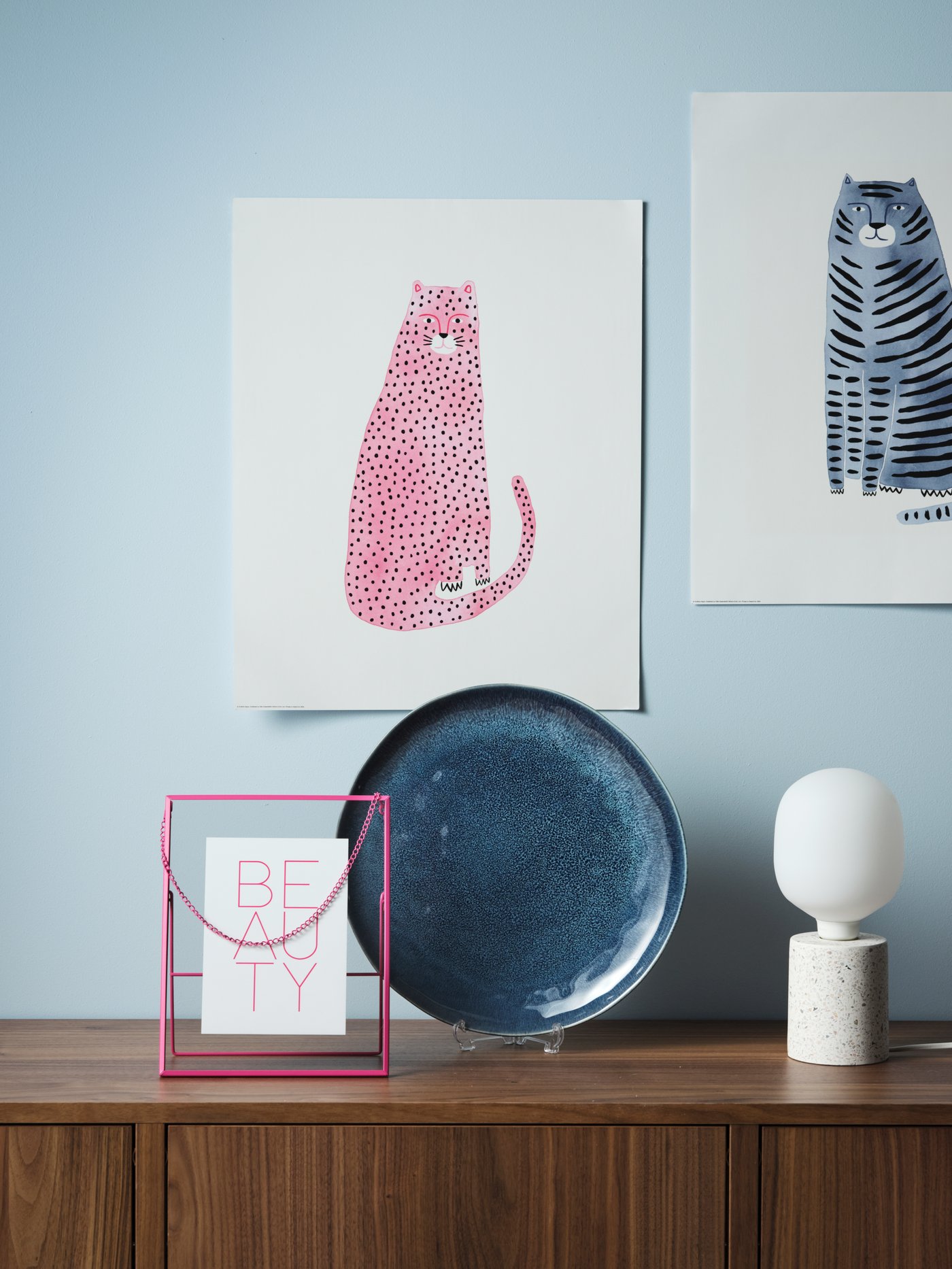 Still life with a pink LERBODA frame, two cat posters, a blue stoneware dish and a table lamp with a decorative light bulb.