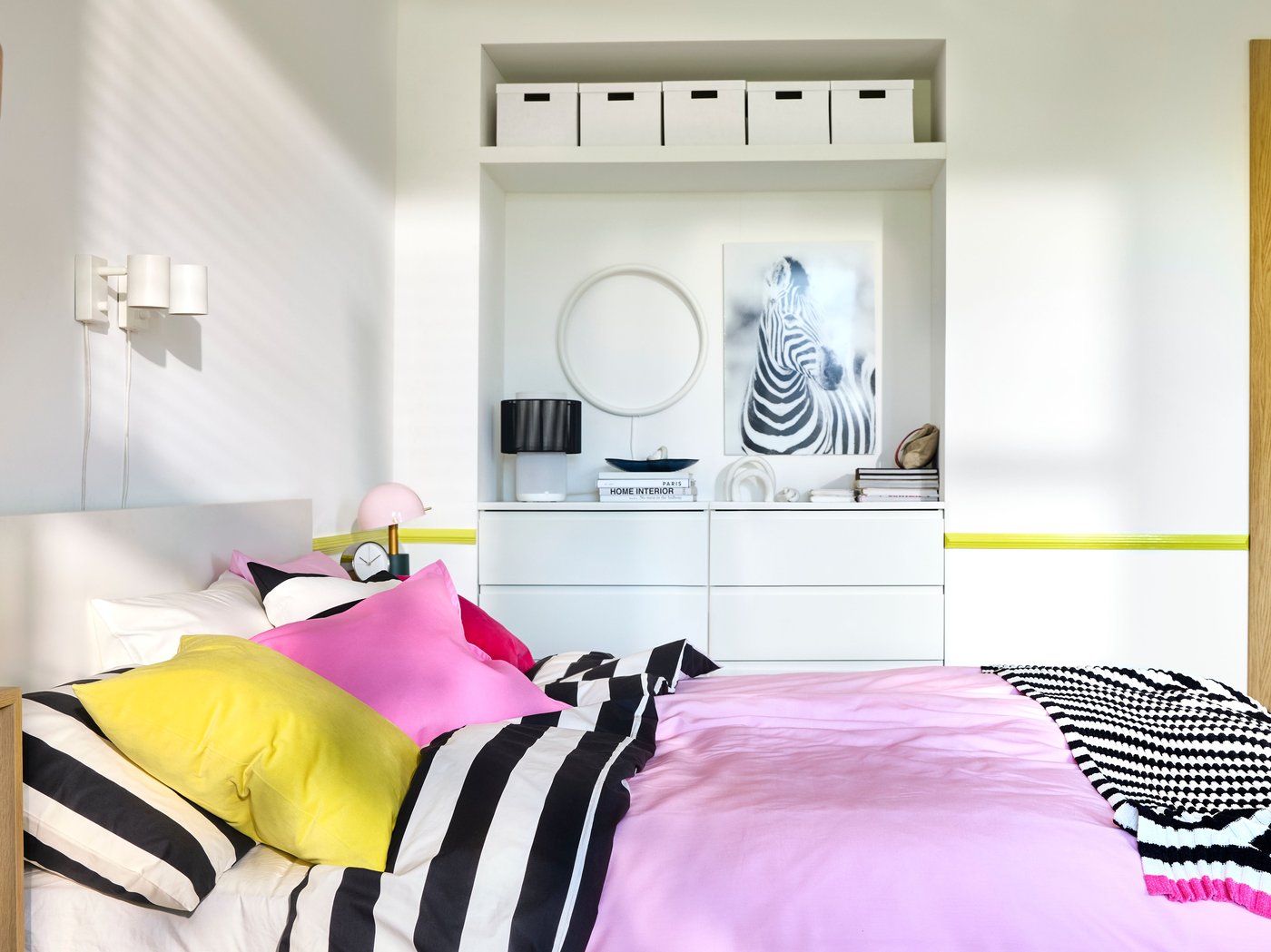 Chambre à coucher aux murs blancs avec rangements intégrés derrière un lit deux places habillé d’un linge de lit rayé en rose vif et noir/blanc.