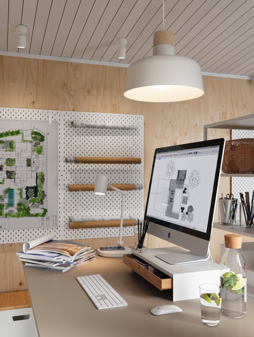 An ELLOVEN monitor stand on top of a desk in a bright studio. A pendant lamp is above and a small work lamp beside it.