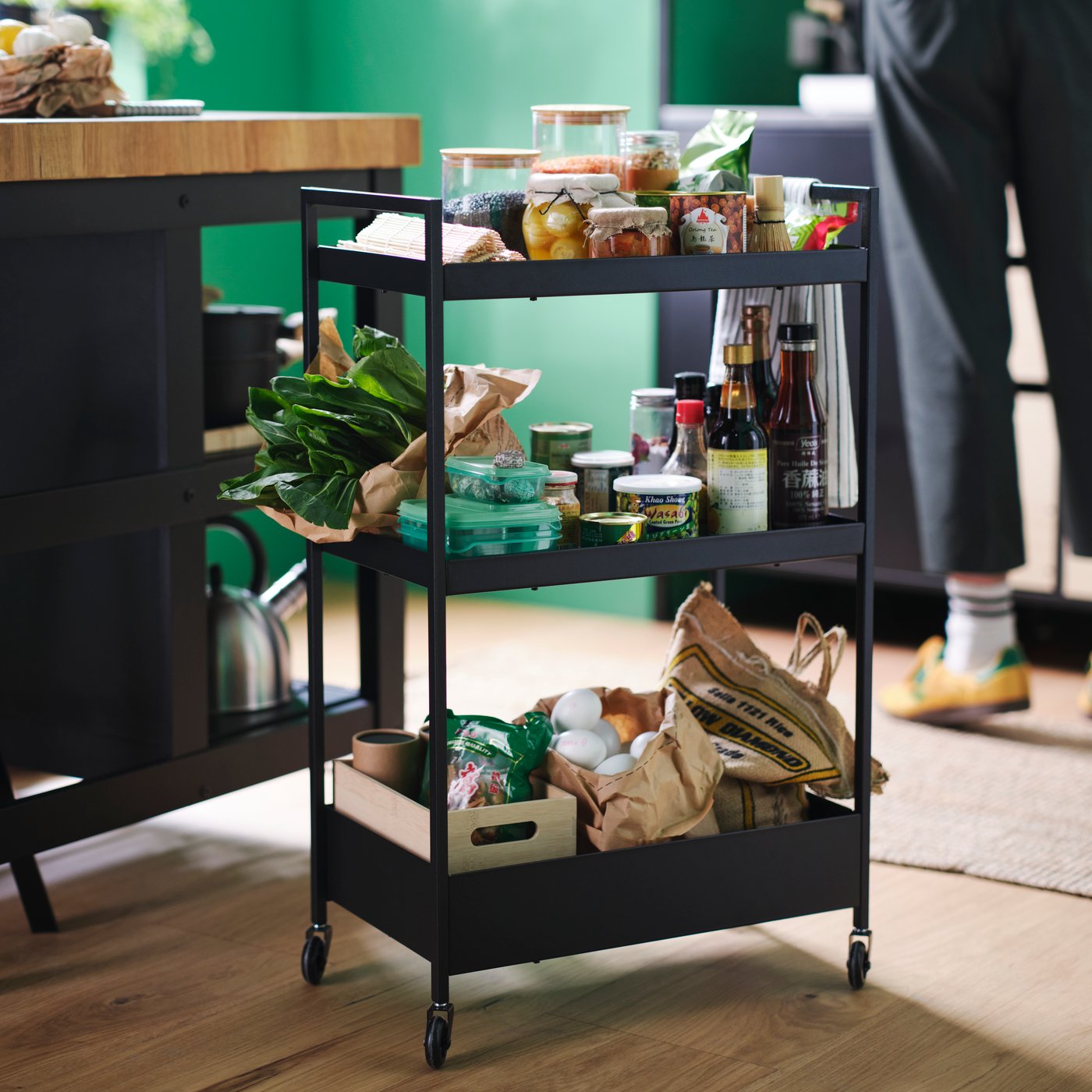 The shelves of a black NISSAFORS trolley are teeming with fresh vegetables, eggs, jars and cans of food, and various bottles.