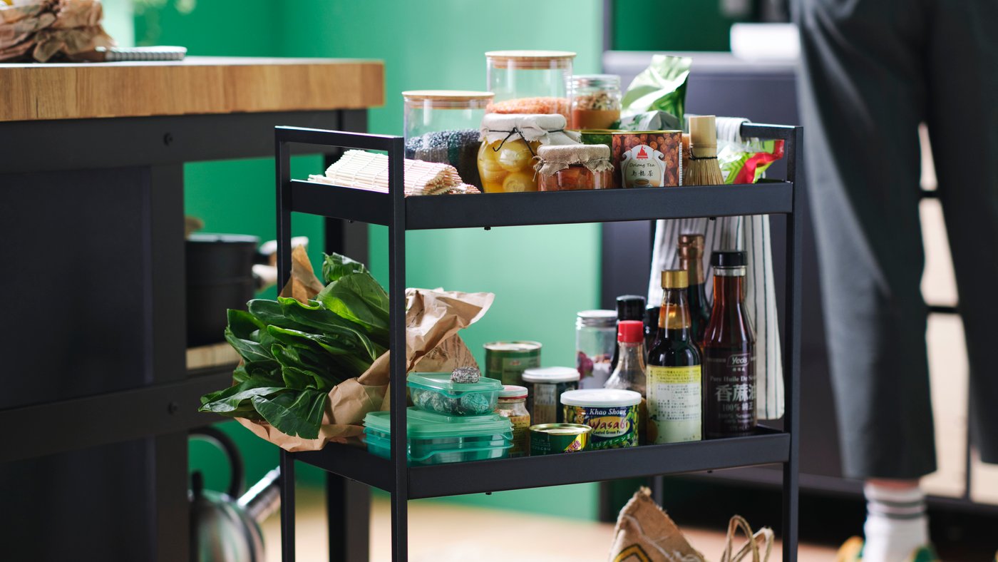 The shelves of a black NISSAFORS trolley are teeming with fresh vegetables, eggs, jars and cans of food, and various bottles.