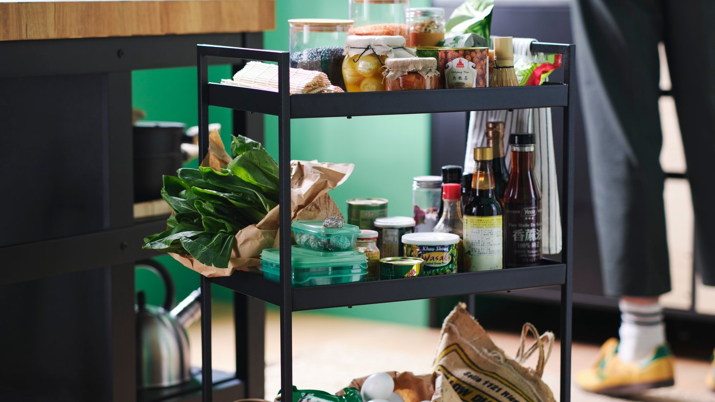 The shelves of a black NISSAFORS trolley are teeming with fresh vegetables, eggs, jars and cans of food, and various bottles.