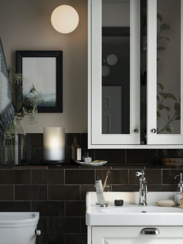A dimly lit bathroom with a white TÄNNFORSEN mirror cabinet with doors and a round FRIHULT lamp on the wall.