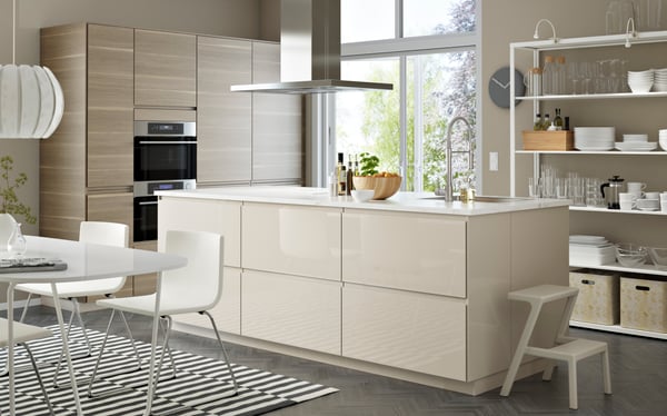 Große IKEA Küchenlösung mit einer Kücheninsel mit einer Abzugshaube. die Küche hat glenzend Besche Fronten und eine Glenzend weiße Arbeitsplatte. auf der Linken Seite befindet sich ein Esstisch mit passenden Stühlen.