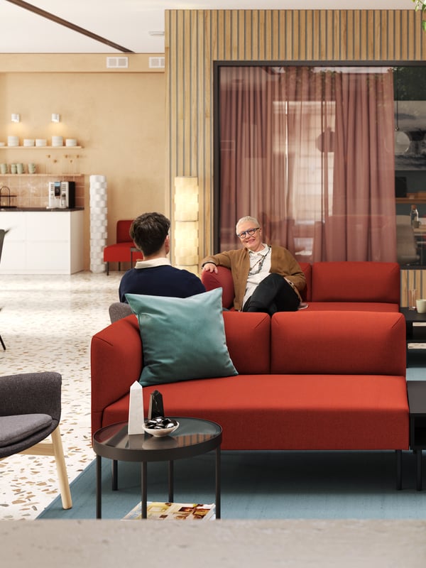 Two people relaxing in a modern, light-filled lobby and meeting room that has two red LILLEHEM modular sofas in the centre.